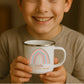 Kind hält weiße Emaille Kindertasse mit silbernem Rand – personalisierte Tasse mit Namen und süßem Motiv
