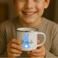 Kind hält weiße Emaille Kindertasse mit silbernem Rand – personalisierte Tasse mit Namen und süßem Motiv